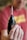 A person is holding a black cordless drill with a metal drill bit attached, prominently displayed in focus. The background is blurred, including a person wearing a red shirt.