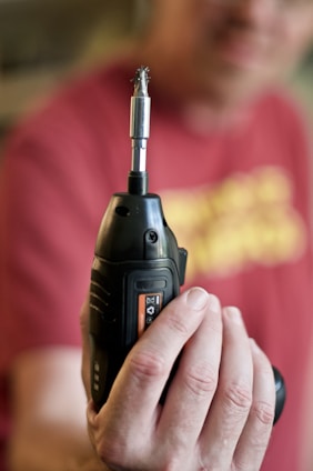 A person is holding a black cordless drill with a metal drill bit attached, prominently displayed in focus. The background is blurred, including a person wearing a red shirt.