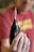 A person is holding a black cordless drill with a metal drill bit attached, prominently displayed in focus. The background is blurred, including a person wearing a red shirt.