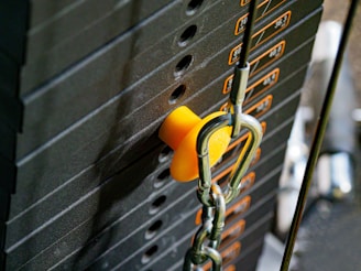 A close-up of a weight stack in a gym, with a focus on the yellow selector pin inserted into the stack. The weights have numbers indicating their mass, and there are chains and cables connected to the equipment.