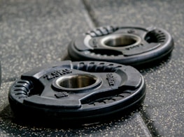 Two black weight plates with the inscription '2.5 kg' and a metal center, placed on a textured gym floor. The lighting highlights their curved, ergonomic design and subtle details.