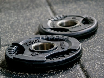 Two black weight plates with the inscription '2.5 kg' and a metal center, placed on a textured gym floor. The lighting highlights their curved, ergonomic design and subtle details.