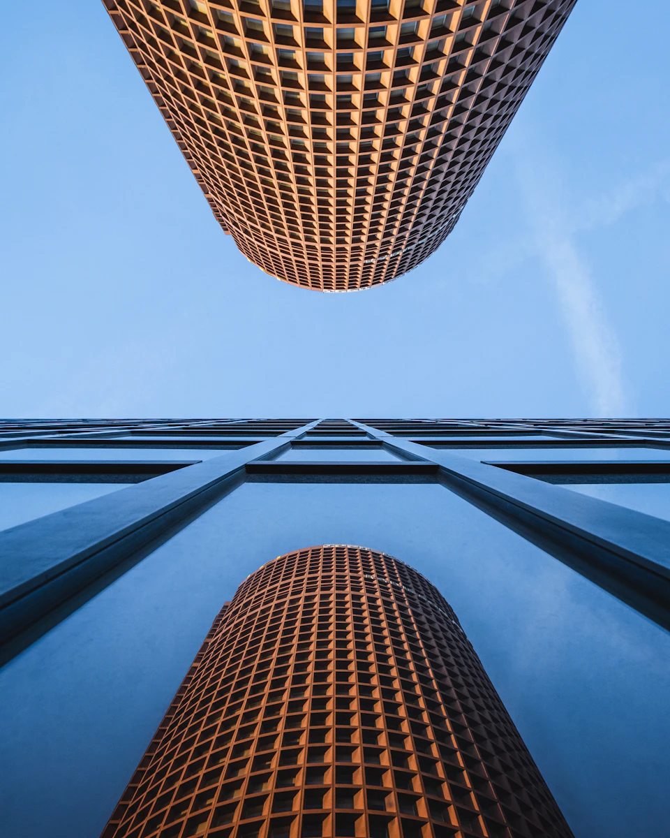 The image shows a tall, cylindrical building with a grid-like pattern on its facade. The perspective is from below, looking up at the structure, which gives it an imposing and majestic appearance. The sky forms the backdrop for the building's reflection in the windows, creating a mirror image that adds depth to the scene.