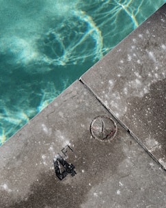 A swimming pool edge with clear aqua-colored water on one side and a concrete surface on the other. The concrete shows markings of '4 FT' indicating the water depth. The surface has areas of wear and discoloration.