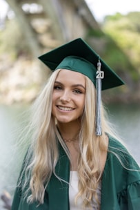 A young woman with long blonde hair is wearing a green graduation cap and gown, standing outdoors with a blurred bridge and greenery in the background. She has a radiant smile and an embellishment with the number '20' hangs from her tassel.