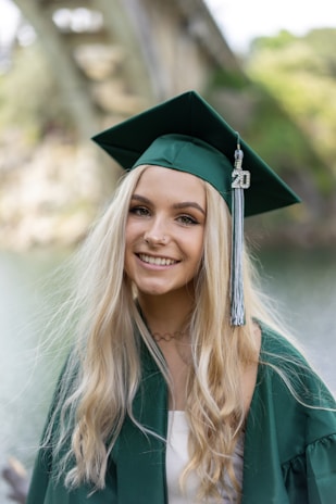 A young woman with long blonde hair is wearing a green graduation cap and gown, standing outdoors with a blurred bridge and greenery in the background. She has a radiant smile and an embellishment with the number '20' hangs from her tassel.