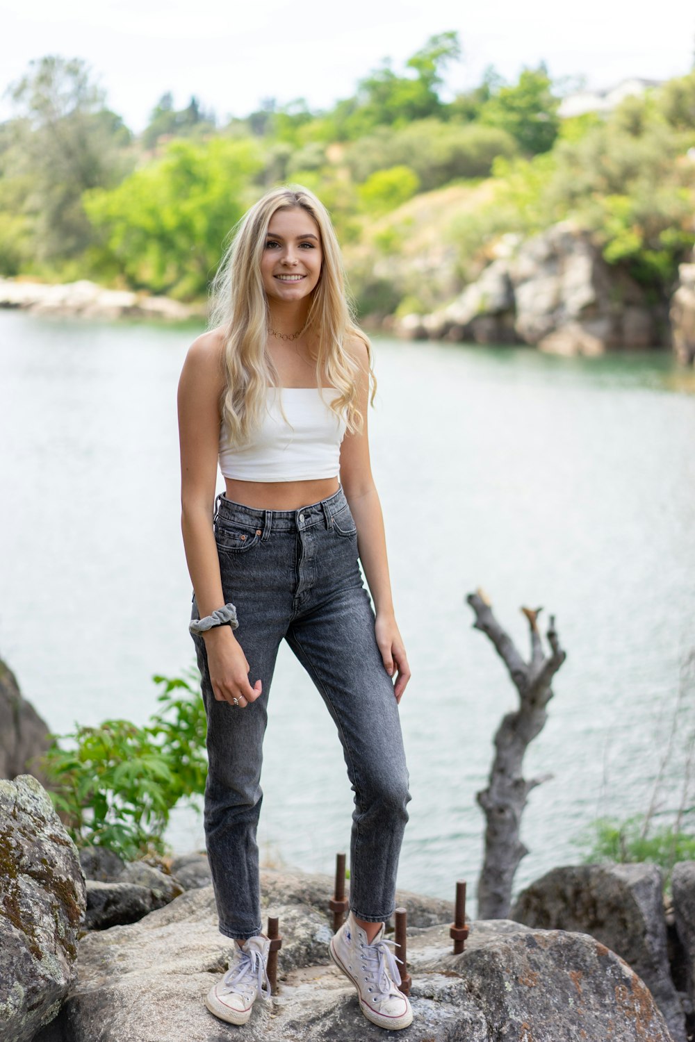 Donna in canotta bianca e jeans blu in denim in piedi sulla roccia vicino allo specchio d'acqua