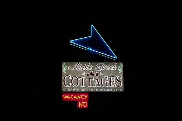 An illuminated sign with a prominent blue neon arrow pointing upwards. Below the arrow, the sign reads 'Apple Creek Cottages' in decorative letters with leaf accents. Additional text mentions 'Suites with Kitchen' and 'Reasonable Rates.' A smaller sign beneath indicates 'Vacancy' in yellow and 'No' in red.