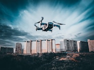 A drone is flying in the foreground above a grassy or dirt area, with a backdrop of tall residential apartment buildings under a cloudy sky. The image captures a dynamic perspective with the drone in focus as it hovers, showcasing the details of the urban landscape.
