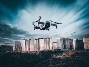 A drone is flying in the foreground above a grassy or dirt area, with a backdrop of tall residential apartment buildings under a cloudy sky. The image captures a dynamic perspective with the drone in focus as it hovers, showcasing the details of the urban landscape.