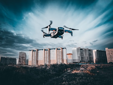 A drone is flying in the foreground above a grassy or dirt area, with a backdrop of tall residential apartment buildings under a cloudy sky. The image captures a dynamic perspective with the drone in focus as it hovers, showcasing the details of the urban landscape.