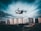 A drone is flying in the foreground above a grassy or dirt area, with a backdrop of tall residential apartment buildings under a cloudy sky. The image captures a dynamic perspective with the drone in focus as it hovers, showcasing the details of the urban landscape.