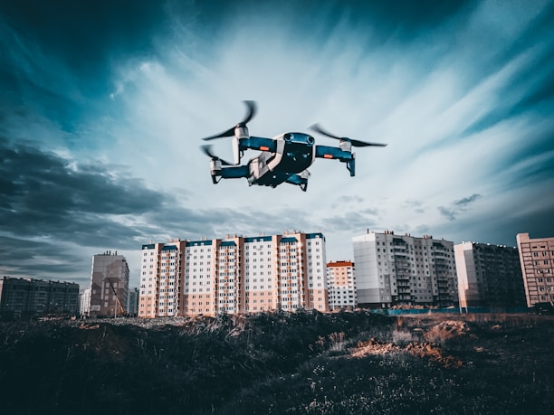 A drone is flying in the foreground above a grassy or dirt area, with a backdrop of tall residential apartment buildings under a cloudy sky. The image captures a dynamic perspective with the drone in focus as it hovers, showcasing the details of the urban landscape.