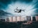 A drone is flying in the foreground above a grassy or dirt area, with a backdrop of tall residential apartment buildings under a cloudy sky. The image captures a dynamic perspective with the drone in focus as it hovers, showcasing the details of the urban landscape.