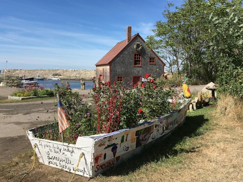 A quaint wooden house with a red roof sits near a waterfront, accompanied by a creatively painted boat filled with vibrant garden flowers. The background includes a stone breakwater and small boats moored on a serene body of water. The scene is surrounded by greenery with a bright blue sky overhead.