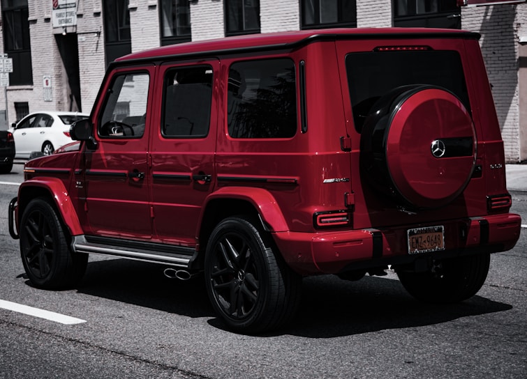 A red luxury SUV parked on a city street, featuring sleek lines and a spare tire mounted on the back. The building in the background has a brick facade with several windows. Other cars are parked along the street, contributing to an urban setting.