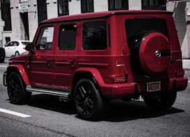 A red luxury SUV parked on a city street, featuring sleek lines and a spare tire mounted on the back. The building in the background has a brick facade with several windows. Other cars are parked along the street, contributing to an urban setting.