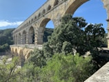 An ancient stone aqueduct with multiple arches spans across a lush, green landscape. The structure is built from large rectangular stones, showcasing impressive engineering. Dense trees and vegetation surround the aqueduct, and a clear blue sky completes the serene setting.
