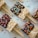 Rectangular crispy rice treats are partially dipped in chocolate and decorated with colorful sprinkles. Each treat is attached to a popsicle stick and laid out on a light-colored surface. Various shapes and colors of sprinkles add a playful touch.
