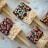 Rectangular crispy rice treats are partially dipped in chocolate and decorated with colorful sprinkles. Each treat is attached to a popsicle stick and laid out on a light-colored surface. Various shapes and colors of sprinkles add a playful touch.