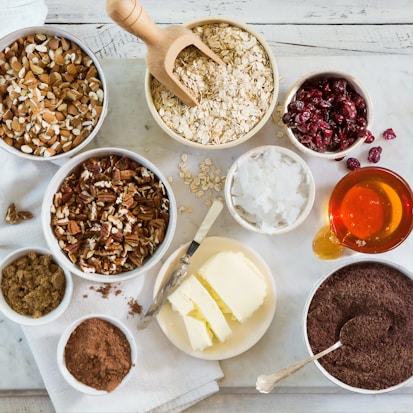 An assortment of ingredients is arranged on a white wooden table, including bowls of almonds, pecans, oats, dried cranberries, coconut flakes, brown sugar, cocoa powder, and grated chocolate. There's a plate with slices of butter accompanied by a knife, and a small pitcher filled with honey.