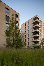 Two modern apartment buildings are surrounded by green foliage and grass. The buildings have brick exteriors with balconies and windows, set against a backdrop of a clear blue sky.