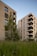 Two modern apartment buildings are surrounded by green foliage and grass. The buildings have brick exteriors with balconies and windows, set against a backdrop of a clear blue sky.