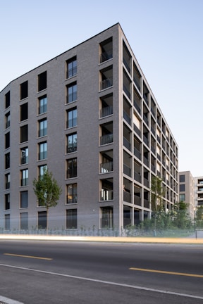 A modern multi-story apartment building with a minimalist architectural style, characterized by its rectangular shape and large windows. The structure is surrounded by greenery including trees and small bushes. The sky is clear, and the image captures a street in the foreground with a trail of light, indicative of a passing vehicle, creating a dynamic contrast against the stillness of the building.