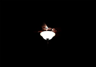 A ceiling light fixture is featured prominently against a dark background. The light emits a soft, warm glow that slightly illuminates the surrounding area. The fixture's design includes metal components surrounding the light source.