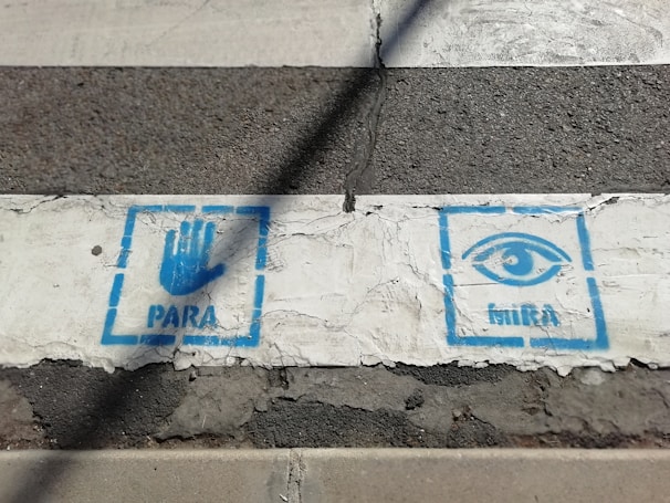 A crosswalk with white paint and black asphalt. There are two blue painted symbols on the white section. On the left, a blue hand with the word 'PARA' below it. On the right, an eye with the word 'MIRA' underneath.
