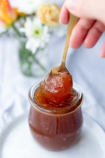 A hand holding a spoon scoops a thick, amber-colored jelly from a glass jar. In the background, blurred flowers in a vase add a touch of color.