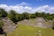 Ancient stone pyramids and structures are surrounded by lush green forest under a bright blue sky with scattered clouds. Stone steps lead up to the main pyramidal structures, set amid a cleared grassy area.