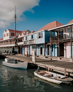 A waterfront scene featuring a row of colorful wooden buildings with a rustic aesthetic, some with signage, along a dock. There are small boats moored in calm water. The sky is clear with a few clouds, and a person is walking on the dock.