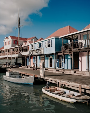 A waterfront scene featuring a row of colorful wooden buildings with a rustic aesthetic, some with signage, along a dock. There are small boats moored in calm water. The sky is clear with a few clouds, and a person is walking on the dock.