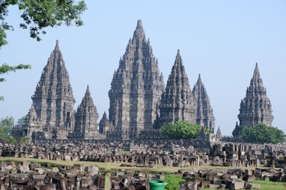 An ancient temple complex featuring multiple intricately carved spires surrounded by stone ruins and lush green foliage. The architecture reflects traditional Southeast Asian design with tall, conical towers and detailed stonework.
