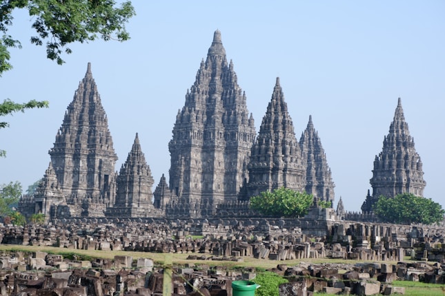 An ancient temple complex featuring multiple intricately carved spires surrounded by stone ruins and lush green foliage. The architecture reflects traditional Southeast Asian design with tall, conical towers and detailed stonework.