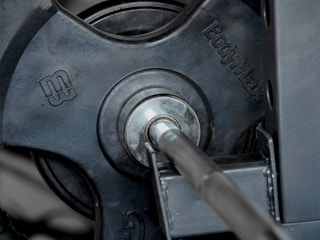 Close-up view of a weight plate attached to a barbell on a gym equipment. The brand name 'BodyMax' is embossed on the plate, which appears to be made of dark metal.