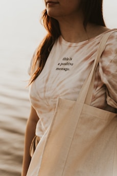 A person is partially visible, wearing a tie-dye T-shirt with the text 'this is a positive message.' They are carrying a beige tote bag, and the background features a soft, blurred view of what appears to be water, suggesting a peaceful outdoor setting.