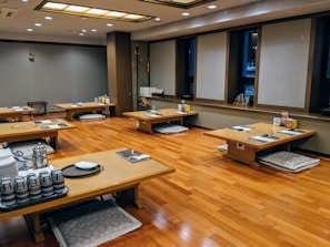 A traditional dining area with low wooden tables arranged in rows. Each table is set with placemats, teacups, and other dining accessories. The floor is made of polished wood, and cushions are placed beneath each table for seating. The walls are decorated with minimalist decor and there are several large windows with simple blinds.