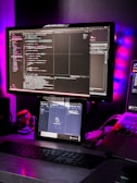 A workspace setup featuring a large monitor displaying code with a dark theme. Below the monitor, a tablet also shows code. A keyboard is placed on the desk, and a laptop is partially visible on the side. Ambient lighting with purple and blue hues creates a modern and tech-focused atmosphere. Various items, including a small figurine, are present on the desk.