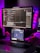 A workspace setup featuring a large monitor displaying code with a dark theme. Below the monitor, a tablet also shows code. A keyboard is placed on the desk, and a laptop is partially visible on the side. Ambient lighting with purple and blue hues creates a modern and tech-focused atmosphere. Various items, including a small figurine, are present on the desk.