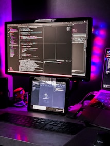 A workspace setup featuring a large monitor displaying code with a dark theme. Below the monitor, a tablet also shows code. A keyboard is placed on the desk, and a laptop is partially visible on the side. Ambient lighting with purple and blue hues creates a modern and tech-focused atmosphere. Various items, including a small figurine, are present on the desk.
