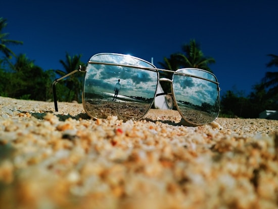 Sunglasses lie on a sandy surface, reflecting a beach scene with a person and scattered clouds against a blue sky.
