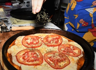 A close-up of a homemade pizza topped with sliced tomatoes and melted cheese, being sprinkled with herbs. The pizza rests on a dark baking tray, and a hand above is adding a finishing touch. Items such as a rolling pin and a colorful apron are visible in the background.