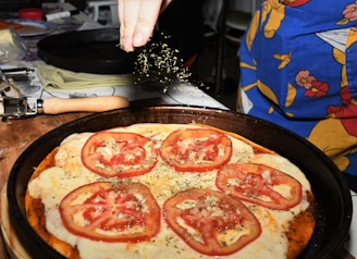 A close-up of a homemade pizza topped with sliced tomatoes and melted cheese, being sprinkled with herbs. The pizza rests on a dark baking tray, and a hand above is adding a finishing touch. Items such as a rolling pin and a colorful apron are visible in the background.