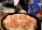 A close-up of a homemade pizza topped with sliced tomatoes and melted cheese, being sprinkled with herbs. The pizza rests on a dark baking tray, and a hand above is adding a finishing touch. Items such as a rolling pin and a colorful apron are visible in the background.