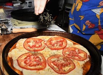 A close-up of a homemade pizza topped with sliced tomatoes and melted cheese, being sprinkled with herbs. The pizza rests on a dark baking tray, and a hand above is adding a finishing touch. Items such as a rolling pin and a colorful apron are visible in the background.