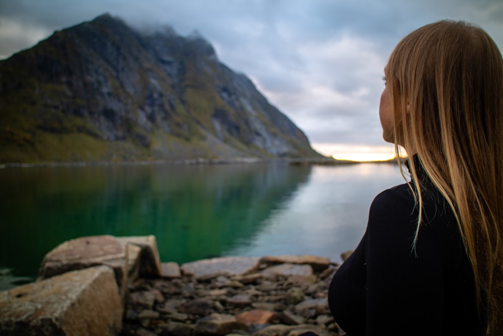 woman in black jacket sitting on rock near lake during daytime