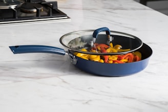A blue frying pan with a glass lid filled with sliced red and yellow bell peppers, placed on a light-colored marble countertop. A stovetop is visible in the background.