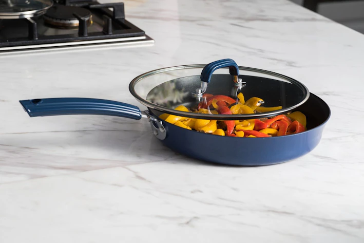 A set of colorful non-stick frying pans arranged on a wooden kitchen countertop with fresh herbs nearby.